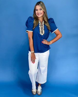 Woman wearing a blue top with white trim and white pants against a blue background