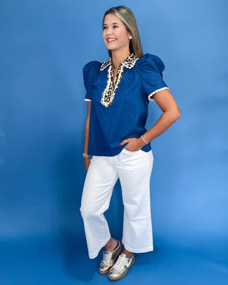 Woman wearing a blue top with leopard print trim and white pants against a blue background