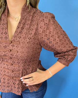 Chocolate brown eyelet peplum top with a V-neckline, decorative bow-and-button front details, and three-quarter puff sleeves.