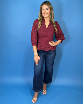 Burgundy eyelet peplum top with a V-neckline, decorative bow-and-button front details, and three-quarter puff sleeves.