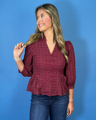 Burgundy eyelet peplum top with a V-neckline, decorative bow-and-button front details, and three-quarter puff sleeves.