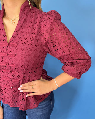 Burgundy eyelet peplum top with a V-neckline, decorative bow-and-button front details, and three-quarter puff sleeves.