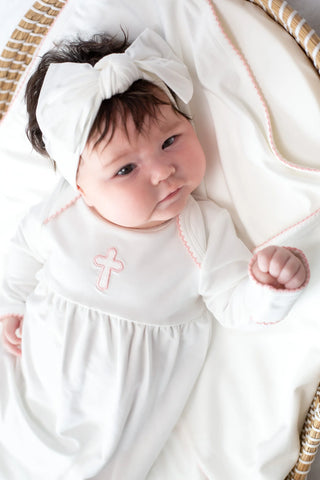 Newborn baby in a white outfit with a cross design, wearing a white headband, lying on a white blanket.