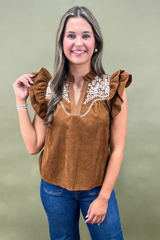 Woman wearing a brown blouse with ruffled sleeves and white embroidery on a green background