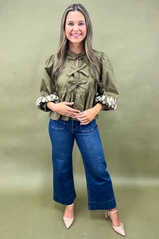 Olive green blouse with bow front detail, puff sleeves, and white embroidered leaf cuffs paired with dark denim wide-leg jeans.