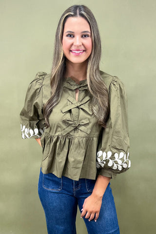 Olive green blouse with bow front detail, puff sleeves, and white embroidered leaf cuffs paired with dark denim wide-leg jeans.