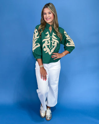 Woman wearing a green blouse with white patterns and white pants against a blue background