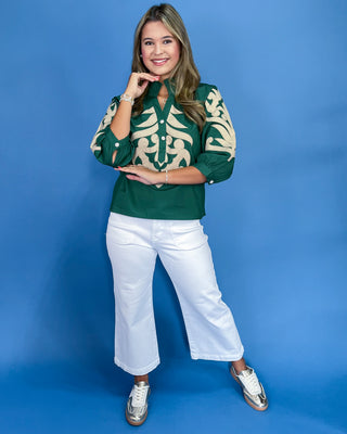 Woman wearing a green and beige patterned shirt with white pants on a blue background