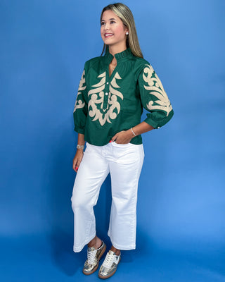 Woman wearing a green blouse with white patterns and white pants against a blue background