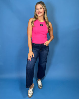 A woman modeling a bright pink sleeveless tank top with a small black logo patch on the chest, paired with dark blue jeans and white sneakers