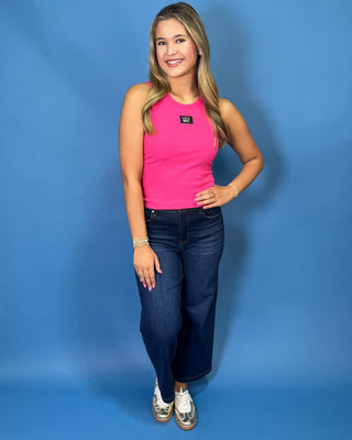 A woman modeling a bright pink sleeveless tank top with a small black logo patch on the chest, paired with dark blue jeans and white sneakers