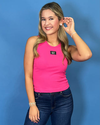 A woman modeling a bright pink sleeveless tank top with a small black logo patch on the chest, paired with dark blue jeans and white sneakers