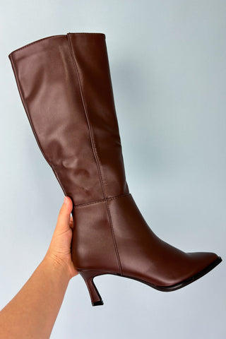 Brown knee-high boot held by a hand against a light gray background