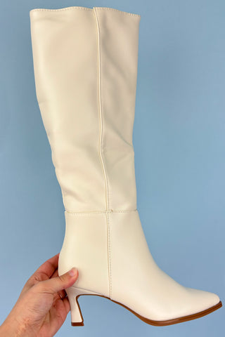 White knee-high boot held by a hand against a light blue background