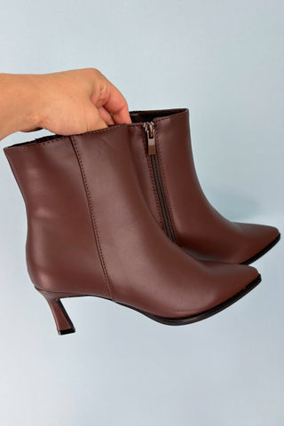 Brown leather ankle boots with a hand holding one boot on a light gray background