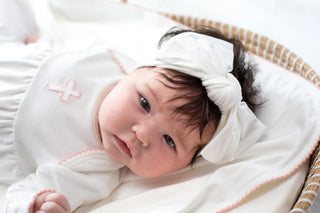 Baby lying on a white blanket with a headband, wearing a white outfit with pink accents.
