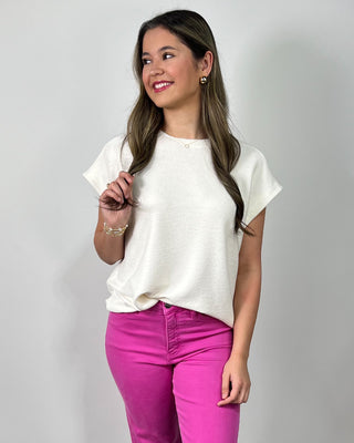 Smiling woman wearing a cream short-sleeve top with bright fuchsia cropped jeans and metallic gold sneakers against a light gray backdrop.