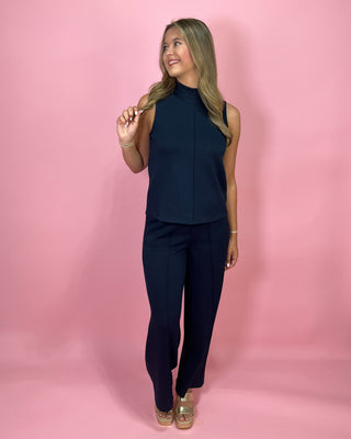 Woman wearing a navy sleeveless top and pants against a pink background