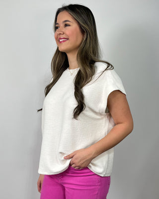 Smiling woman wearing a cream short-sleeve top with bright fuchsia cropped jeans and metallic gold sneakers against a light gray backdrop.