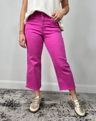 Close-up of bright fuchsia high-rise cropped jeans with a raw hem, styled with a cream top and metallic gold sneakers.