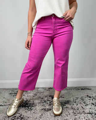 Close-up of bright fuchsia high-rise cropped jeans with a raw hem, styled with a cream top and metallic gold sneakers.