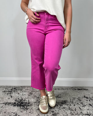 Close-up of bright fuchsia high-rise cropped jeans with a raw hem, styled with a cream top and metallic gold sneakers.