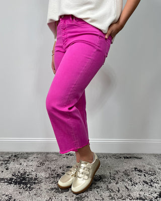 Close-up of bright fuchsia high-rise cropped jeans with a raw hem, styled with a cream top and metallic gold sneakers.