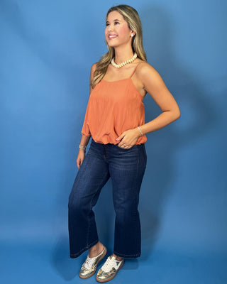 A woman modeling a sleeveless orange top with dark blue jeans and metallic sneakers