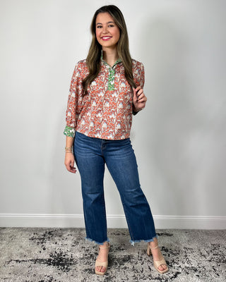 Woman wearing a red and cream floral print button-front blouse with green contrast trim and cuff details, styled with medium-wash cropped jeans and nude heeled sandals, posing against a light gray backdrop.