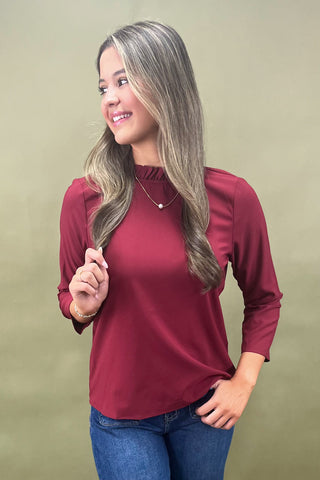 Woman wearing a burgundy long-sleeve top and blue jeans against a beige background