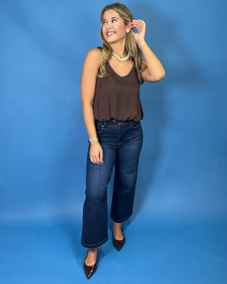 A woman modeling a brown sleeveless top and dark blue wide-leg jeans against a solid blue studio backdrop.