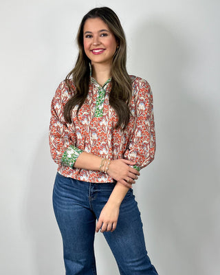 Woman wearing a red and cream floral print button-front blouse with green contrast trim and cuff details, styled with medium-wash cropped jeans and nude heeled sandals, posing against a light gray backdrop.