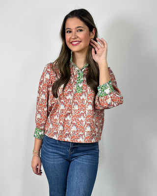 Woman wearing a red and cream floral print button-front blouse with green contrast trim and cuff details, styled with medium-wash cropped jeans and nude heeled sandals, posing against a light gray backdrop.