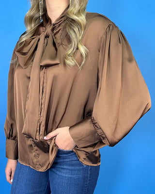 Chocolate brown satin blouse with a statement tie-neck bow, long balloon sleeves, button cuffs, and an elegant relaxed silhouette.
