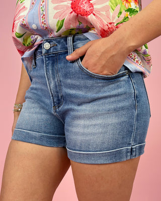 Person wearing blue denim shorts with a floral shirt on a pink background