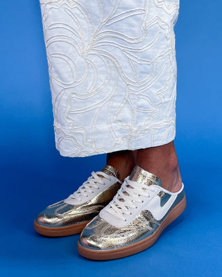 Gold and white sneakers worn with white pants on a blue background