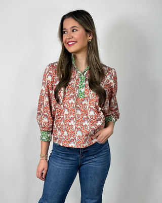Woman wearing a red and cream floral print button-front blouse with green contrast trim and cuff details, styled with medium-wash cropped jeans and nude heeled sandals, posing against a light gray backdrop.