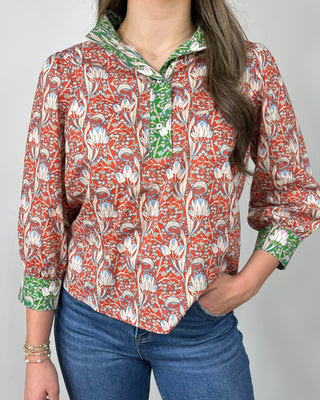 Woman wearing a red and cream floral print button-front blouse with green contrast trim and cuff details, styled with medium-wash cropped jeans and nude heeled sandals, posing against a light gray backdrop.