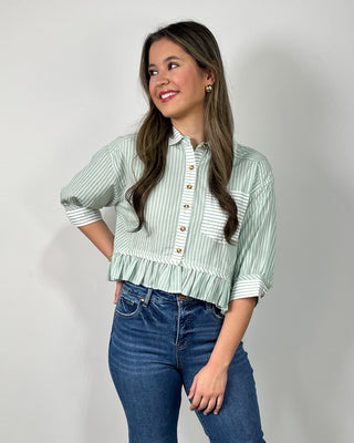Woman wearing a sage green and white striped button-front peplum blouse with three-quarter sleeves, styled with medium-wash cropped flare jeans and nude heeled sandals, posing against a light gray backdrop.