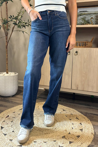 Close-up of a woman wearing high-waisted dark blue straight-leg jeans with a slightly faded wash and raw hems. She pairs them with a white sleeveless knit top featuring black trim and white sneakers, standing on a woven circular rug with a potted plant and wooden shelving in the background.