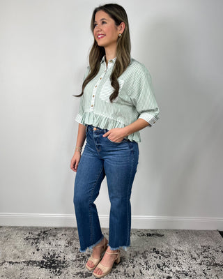 Woman wearing a sage green and white striped button-front peplum blouse with three-quarter sleeves, styled with medium-wash cropped flare jeans and nude heeled sandals, posing against a light gray backdrop.