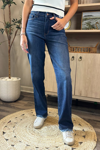 Close-up of a woman wearing high-waisted dark blue straight-leg jeans with a slightly faded wash and raw hems. She pairs them with a white sleeveless knit top featuring black trim and white sneakers, standing on a woven circular rug with a potted plant and wooden shelving in the background.