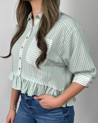 Woman wearing a sage green and white striped button-front peplum blouse with three-quarter sleeves, styled with medium-wash cropped flare jeans and nude heeled sandals, posing against a light gray backdrop.