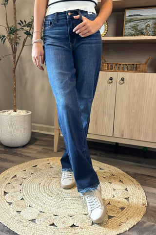 Close-up of a woman wearing high-waisted dark blue straight-leg jeans with a slightly faded wash and raw hems. She pairs them with a white sleeveless knit top featuring black trim and white sneakers, standing on a woven circular rug with a potted plant and wooden shelving in the background.