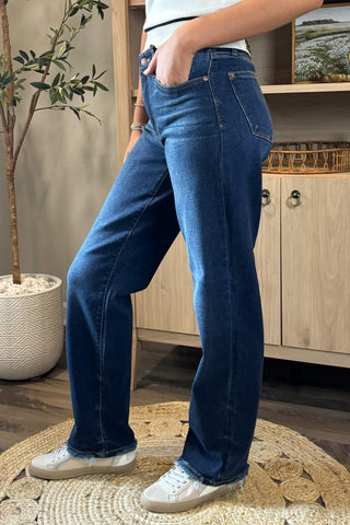 Close-up of a woman wearing high-waisted dark blue straight-leg jeans with a slightly faded wash and raw hems. She pairs them with a white sleeveless knit top featuring black trim and white sneakers, standing on a woven circular rug with a potted plant and wooden shelving in the background.