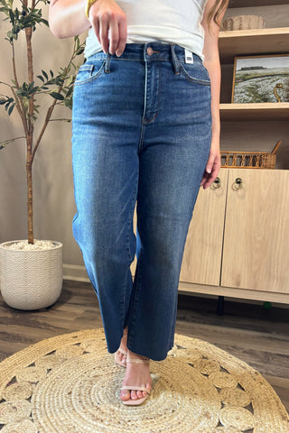 Close-up of a woman wearing high-waisted medium-wash cropped wide-leg jeans. She pairs them with a white ribbed tank top and nude block-heel sandals, standing on a woven circular rug with a potted plant and wooden shelving in the background.