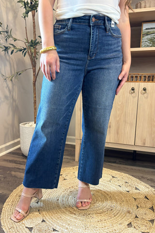 Close-up of a woman wearing high-waisted medium-wash cropped wide-leg jeans. She pairs them with a white ribbed tank top and nude block-heel sandals, standing on a woven circular rug with a potted plant and wooden shelving in the background.