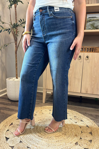 Close-up of a woman wearing high-waisted medium-wash cropped wide-leg jeans. She pairs them with a white ribbed tank top and nude block-heel sandals, standing on a woven circular rug with a potted plant and wooden shelving in the background.