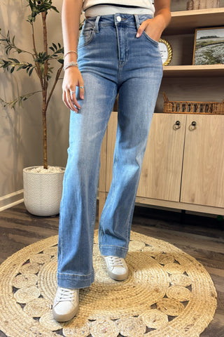 A close-up view of a woman wearing light-wash high-waisted jeans paired with a white sleeveless knit top. She stands indoors on a woven circular rug, wearing white sneakers, with part of a potted plant and wooden shelving visible in the background.