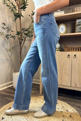 A close-up view of a woman wearing light-wash high-waisted jeans paired with a white sleeveless knit top. She stands indoors on a woven circular rug, wearing white sneakers, with part of a potted plant and wooden shelving visible in the background.